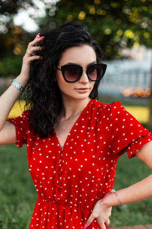 Pretty curly woman with fashion sunglasses and watch in red dress walking outdoors