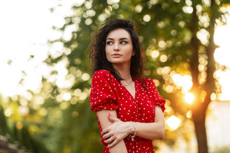Cute curly fashion girl with beautiful face in a summer retro red dress walks in the park at sunsetの写真素材