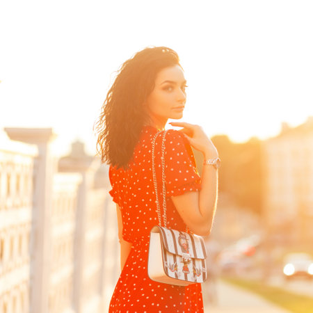 Beautiful young curly woman in vintage red dress with fashion handbag walks on the street at orange sunset and looks at the cameraの写真素材