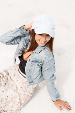 Beautiful young happy woman in fashionable denim clothes with a cap, vintage skirt and black tank top sits on the white sand and rests on summer vacationの写真素材