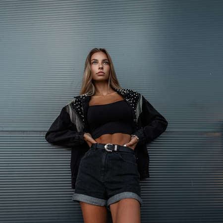 Stylish young girl model with fashion denim jacket in black jeans shorts stands near a black wall...