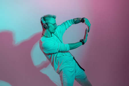 Stylish fashionable handsome man hipster listening to music on headphones and dancing in studio with pink and blue lightの写真素材