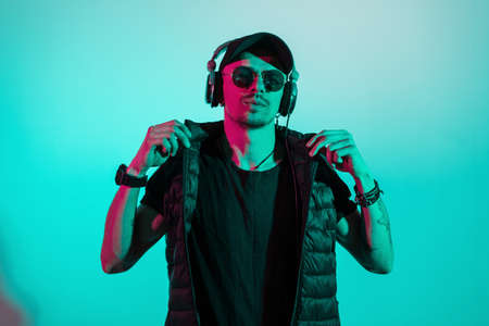 Fashionable handsome hipster man with sunglasses and headphones in stylish black clothes with cap, vest and t-shirt listening to music in studio with creative colored pink and cyan lightの写真素材