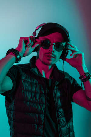 Hipster man with sunglasses and cap listen to music in headphones in studio with colorful pink and...