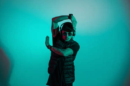 Stylish cool dancer man with black fashion outwear listening to music and dance in colorful studio...