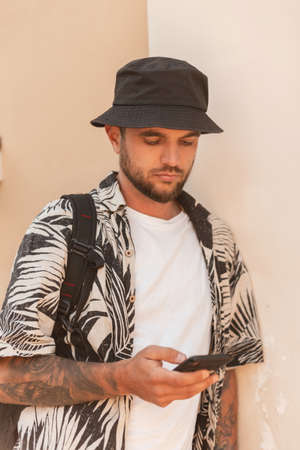 Fashionable handsome hipster man in Hawaiian clothes with a backpack and phone standing near a wall...