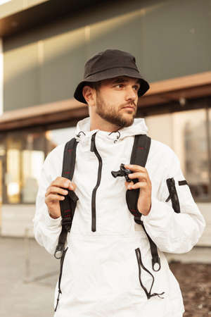 Hipster handsome man model with bucket hat in white sporty jacket with backpack walks on the street...