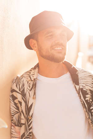 Happy handsome hipster man with smile in fashion shirt with palm trees print and black bucket hat at sunsetの写真素材