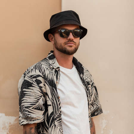 Handsome stylish hipster man with beard, sunglasses and bucket hat in fashion summer outwear with beach shirt near the wall on the streetの写真素材