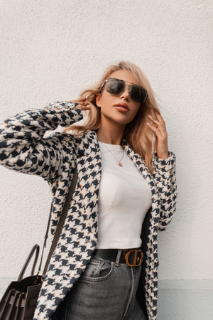 beautiful young girl with glasses in a fashionable coat and a white T-shirt with a bag stands near a wall on the streetの写真素材