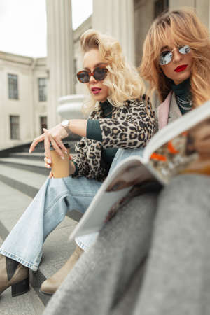 Two fashion beautiful blondes and a redheaded girl in fancy clothes are sitting in town, drinking...