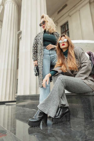 Two glamorous young beautiful girls in fashionable autumn clothes near a vintage building with a columnの写真素材