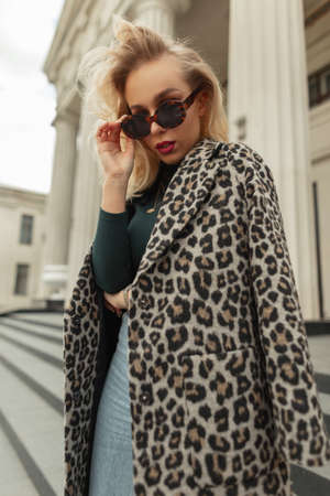 Stylish beautiful young blonde woman puts on fashion sunglasses in trendy clothes with a leopard coat walks in the city near a vintage buildingの写真素材