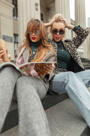 Glamorous young girls in fashionable clothes sit and read a magazine in the cityの写真素材