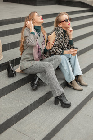 Fashionable happy two girls in trendy spring outfit with sunglasses, leopard jacket, jeans, suit and shoes sits and rest on steps in the city and drinks coffeeの写真素材