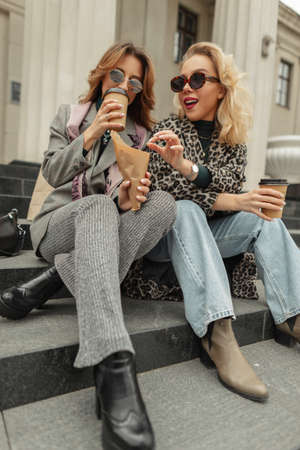 Happy beautiful young girls in fashionable clothes sit on the steps, drinking coffee and eating sweets in the city. travel conceptの写真素材