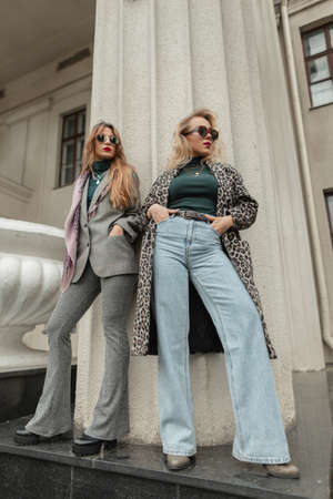 Beautiful young fashionable girls in stylish vintage clothes with coats, high waisted bell-bottom jeans and suits stand near retro columns in the cityの写真素材