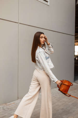 Fashionable pretty woman in stylish casual denim clothes with a jacket and a handbag walks on the street near a gray wallの写真素材