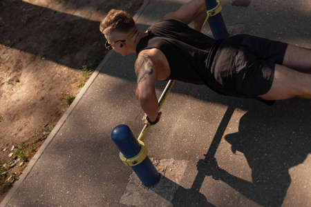 Young handsome sports man with headphones in black sportswear workout and Push-ups on asphalt with metal pipes at sunset. CrossFit and power conceptの写真素材