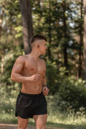 Athlete guy with muscular body runs in the park and listens to music in wireless headphones. Sport...