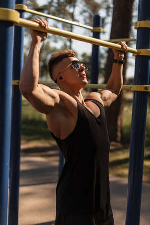 Hipster handsome muscular sports man with headphones and sunglasses in a black tank top pulls himself up on the bar and workout in the park. healthy lifestyleの写真素材