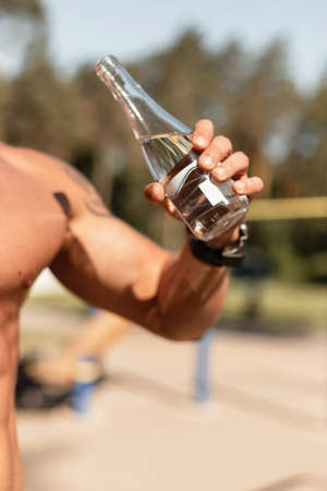 Young man athlete trains and drinks water outdoors. Male muscular arm holding a glass water bottleの写真素材