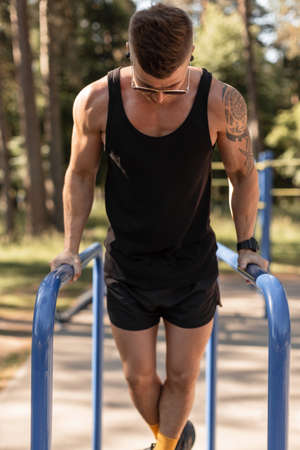 muscular sports man in a black tank top t-shirt and shorts trains and does bars exercises in the...