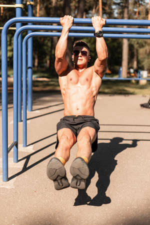 Sports handsome man athlete with muscular naked torso hangs and does exercises on the horizontal bar...
