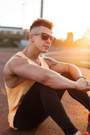 Fashion young hipster fitness man with sunglasses in sportswear sits at stadium at sunsetの写真素材