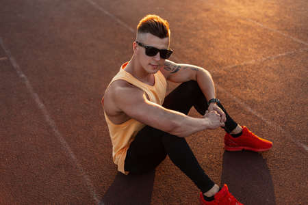 Fitness handsome man athlete with fashion sunglasses in sportswear with red shoes sits and rest on...
