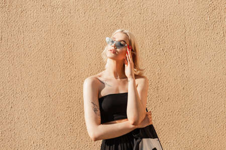 Beautiful fashionable young girl model with short blonde hair in a black top, skirt and sunglasses with cloth bag on the street on sunny dayの写真素材