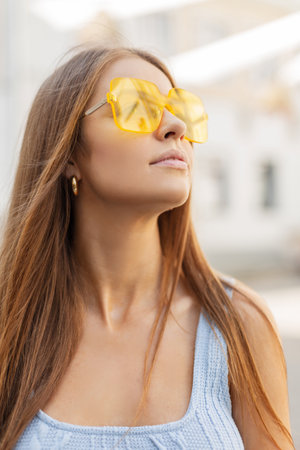 Fresh portrait of a beautiful young girl in a fashionable bright summer outfit with cool yellow sunglasses and a blue knitted tank top is walking in the cityの写真素材