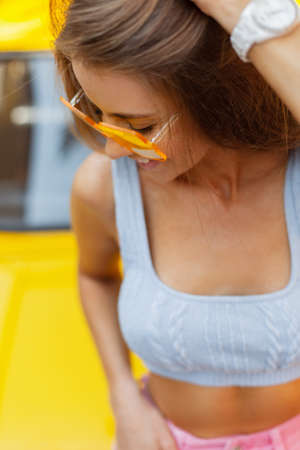 Funny beautiful fashionable girl with a smile in fashion bright colored clothes with a knitted blouse and pink jeans with cool glasses walking in the city near a vintage car on a yellow backgroundの写真素材