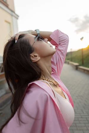 Trendy beautiful model woman with vintage sunglasses and golden chain in pink shirt and top walks and enjoy outdoors at sunset. Female style spring summer look clothesの写真素材
