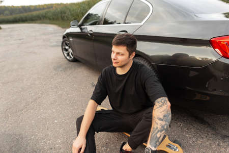 Fashionable hipster man in black T-shirt with tattoo on arm sits by car on the road outside of city