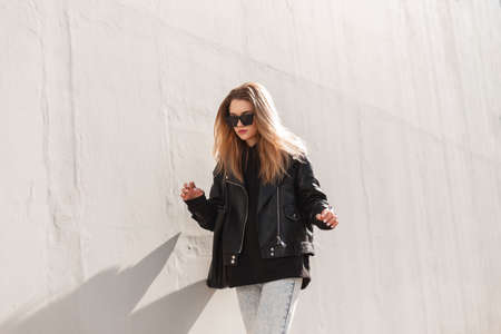 Fashionable hipster woman model with sunglasses in trendy leather rock jacket, hoodie and bag walks...