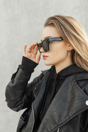 Fashion woman hipster with trendy leather black jacket with black stylish hoodie puts on a cool sunglasses on a gray concrete background. Urban female rock style clothesの写真素材