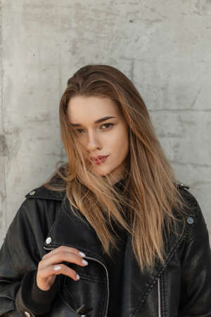 Fashionable urban portrait of young beautiful woman with hairstyle in black fashion leather rock jacket with bag stands near a gray concrete wall on the streetの写真素材
