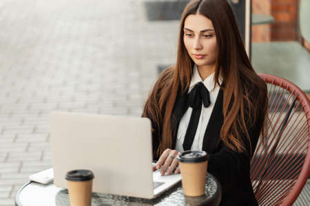 Beautiful professional ht manager woman in a fashion business suit working on a laptop and drinking coffee in a summer cafe on the streetの写真素材