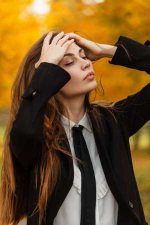 Stylish beautiful young girl in fashionable business clothes enjoying a walk in an autumn park and fixing her hairの写真素材