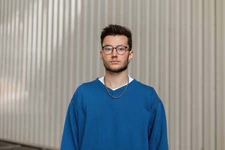Fashionable young stylish hipster man with an earring and glasses in a trendy blue sweater on a gray...