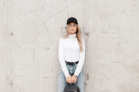 Stylish beautiful young woman with glasses, wearing fashionable clothes with a cap, white sweater...
