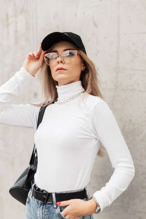 Stylish beautiful young woman with glasses, wearing a fashionable black cap and a white sweater with a backpack stands near a gray concrete wall in the cityの写真素材