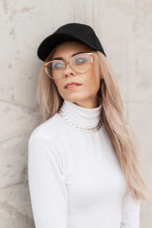 Pretty young woman with glasses and a black cap in a white sweatshirt with a silver chain on the street near a gray concrete wallの写真素材