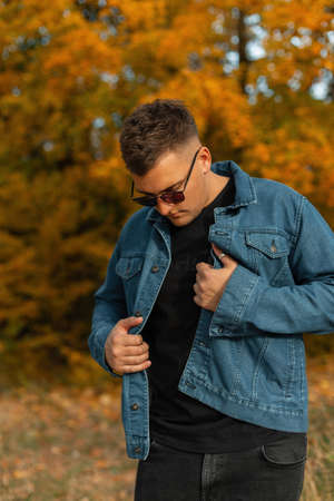 Beautiful stylish hipster man with hairstyle and vintage sunglasses in a fashionable denim shirt and black T-shirt walking in the park with yellow fall foliageの写真素材