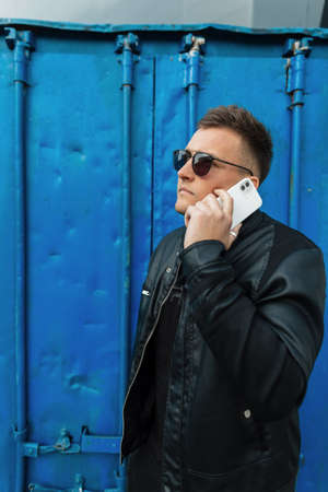 Fashionable handsome hipster man in black clothes with glasses talking on the phone on a blue metallic background in the cityの写真素材