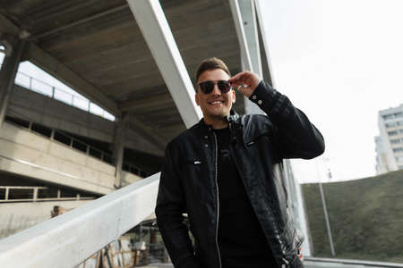 Happiness handsome fashion man hipster model in black leather jacket puts on sunglasses and walks in the cityの写真素材