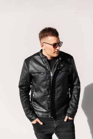 Stylish young hipster guy in a fashion leather jacket and cool sunglasses on the street near the gray wallの写真素材