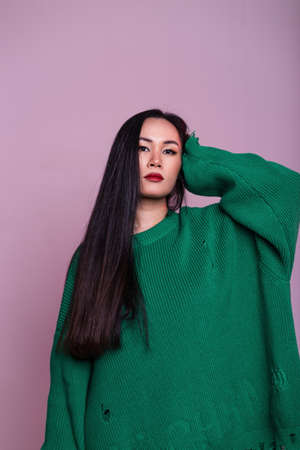 Beautiful glamorous Japanese girl in fashionable clothes with a torn green sweater posing on a pink background in the studioの写真素材