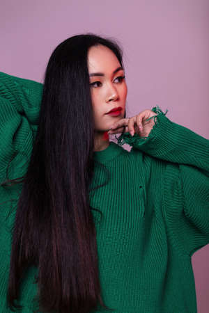 Fashionable beautiful woman model with long healthy hair in stylish knitted green sweater on pink background in studioの写真素材